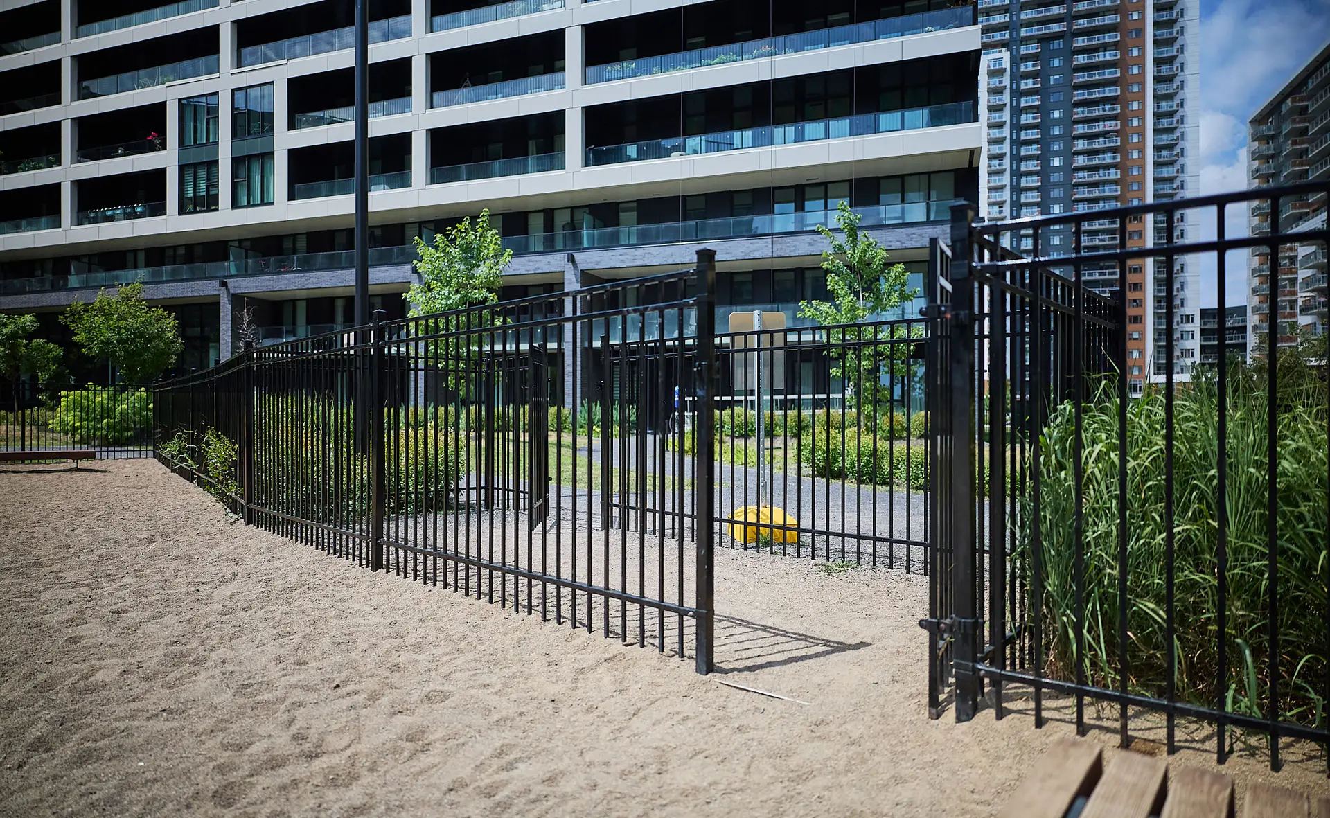 Inter Clôtures Sentinelle A black metal gate stands open in a fence surrounding a sandy area near modern apartment buildings with greenery and trees visible. Fence Installation Quebec