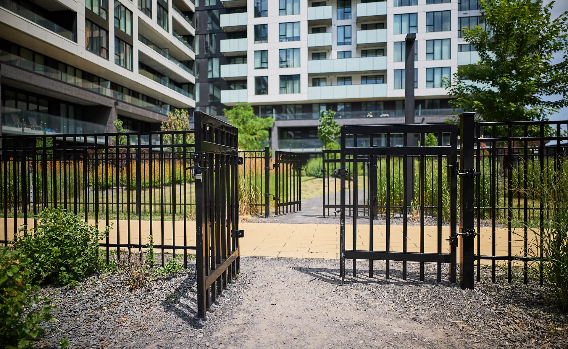 Inter Clôtures Sentinelle Open black metal gate leading to a paved path, surrounded by greenery, with a modern apartment building in the background. Fence Installation Quebec