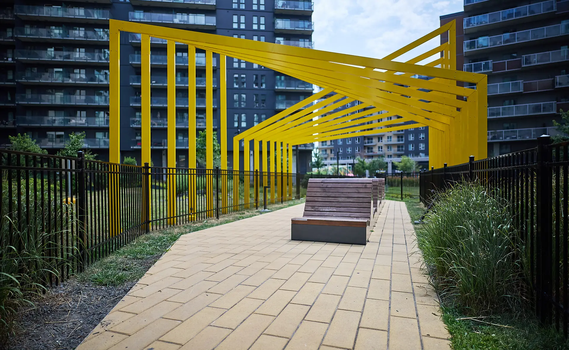 Inter Clôtures Sentinelle A modern park walkway with yellow metal beams, wooden benches, brick path, and fenced grass, surrounded by tall apartment buildings. Fence Installation Quebec