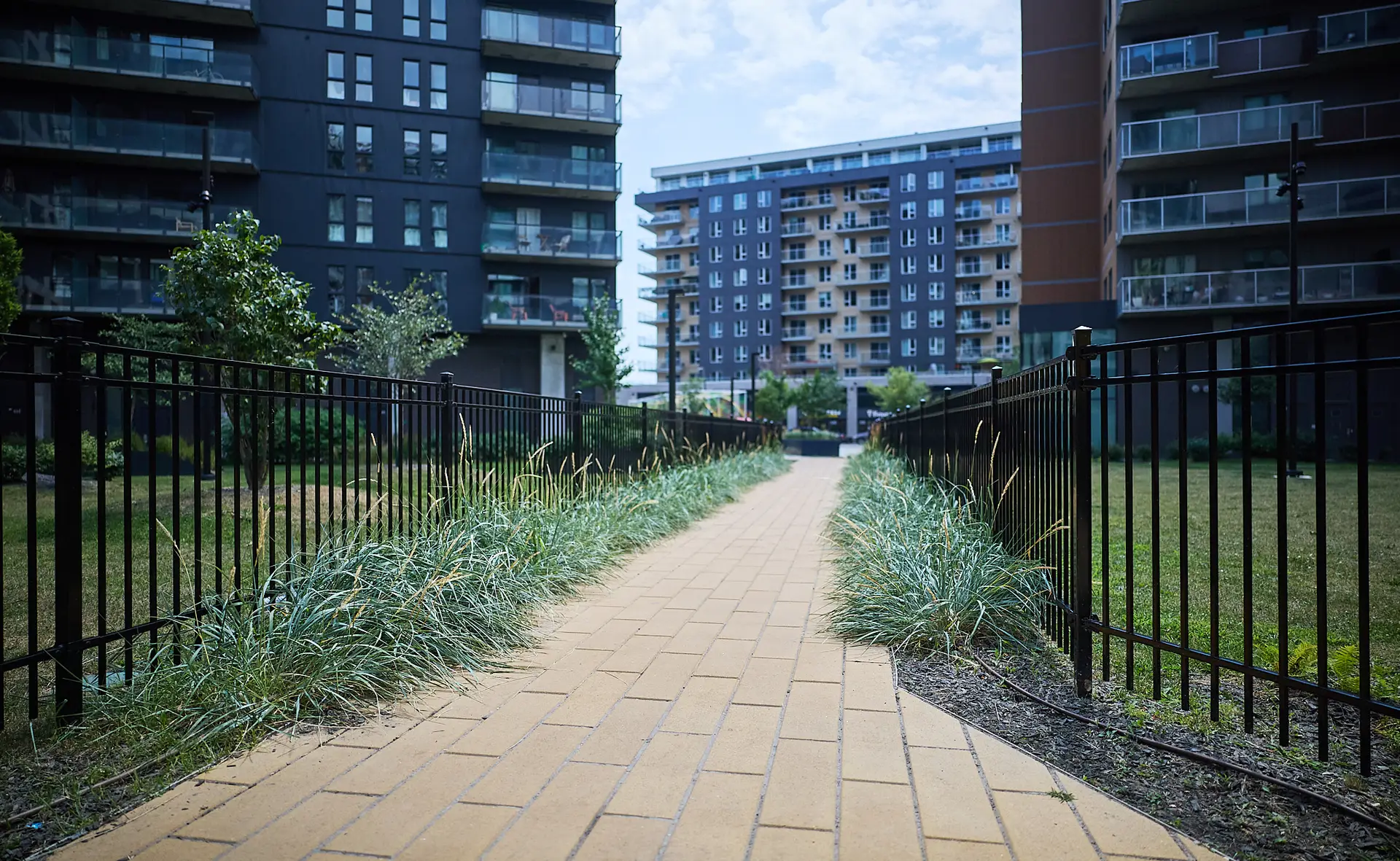 Inter Clôtures Sentinelle A paved walkway with metal railings runs through a grassy courtyard, surrounded by modern apartment buildings under a partly cloudy sky. Fence Installation Quebec