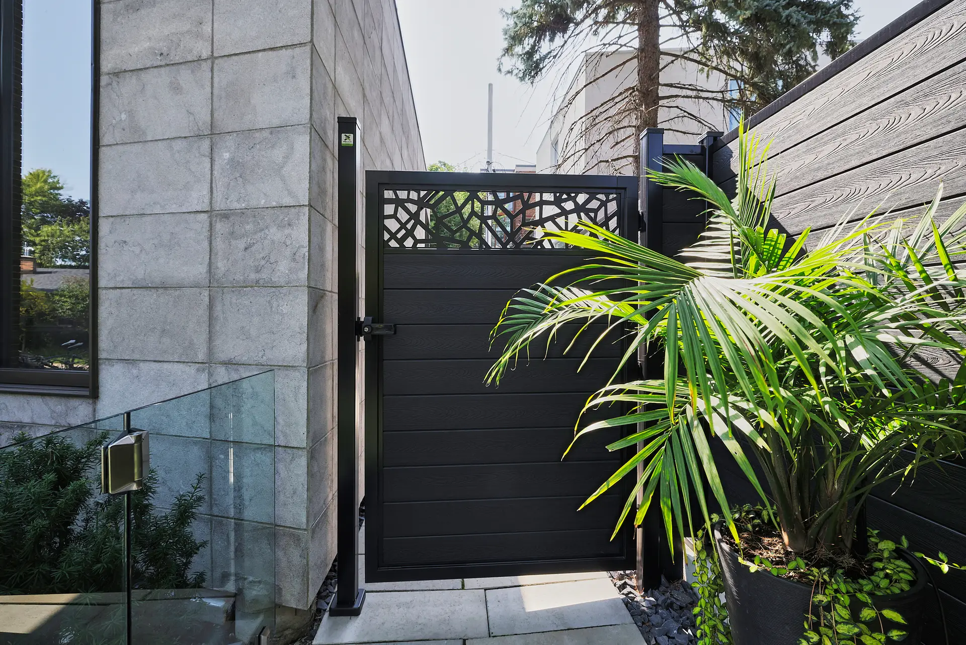 Inter Clôtures Sentinelle A modern black gate with a decorative top panel stands between a tiled building and a wooden fence, next to a green potted palm plant on a stone patio. Fence Installation Quebec