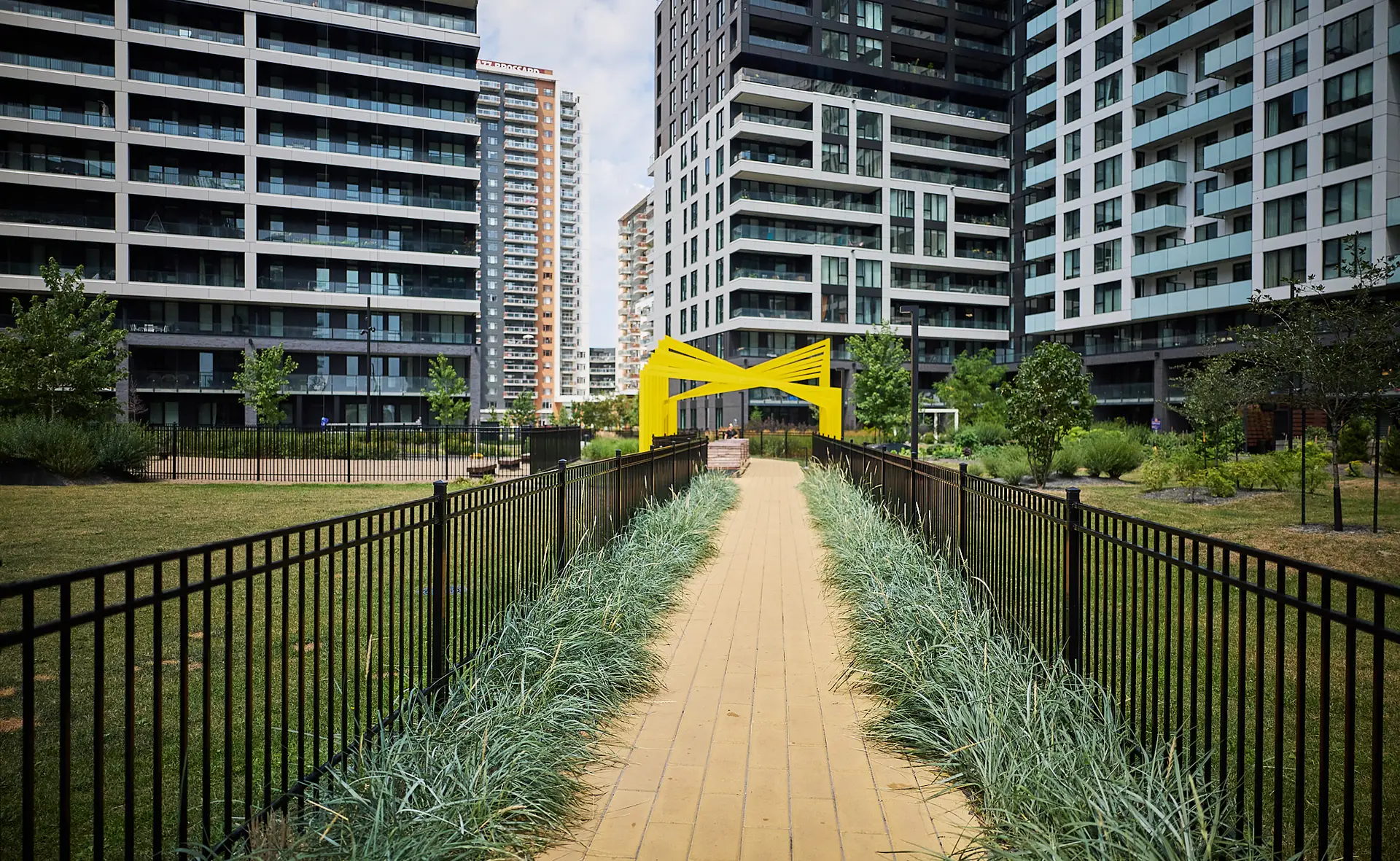 Inter Clôtures Sentinelle A yellow walkway with greenery on both sides leads to a modern yellow sculpture, surrounded by tall residential buildings and landscaped gardens. Fence Installation Quebec