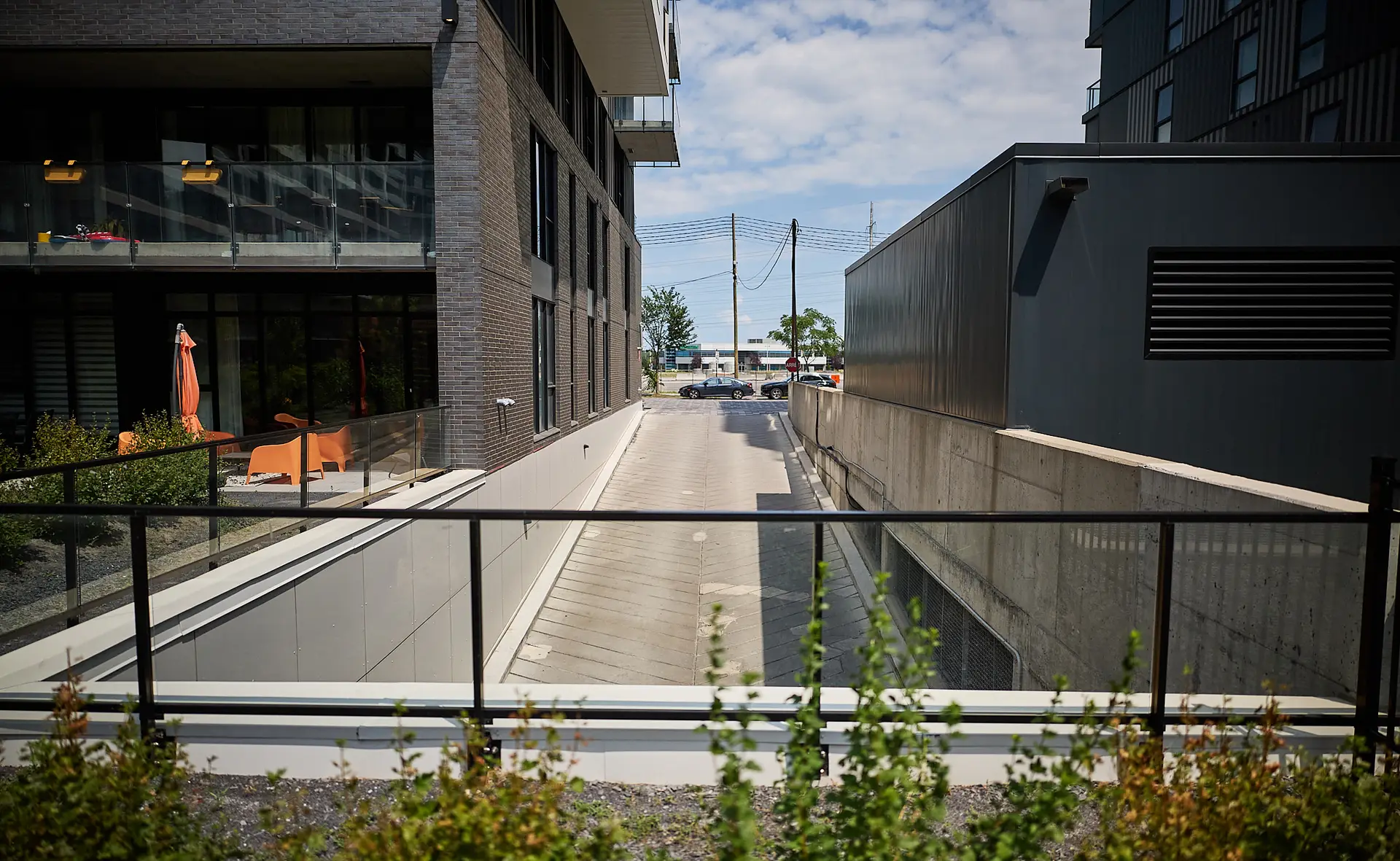 Inter Clôtures Sentinelle A concrete driveway between two modern buildings leads to a street with parked cars. Some greenery is in the foreground, and balconies with orange chairs are visible on the left. Fence Installation Quebec