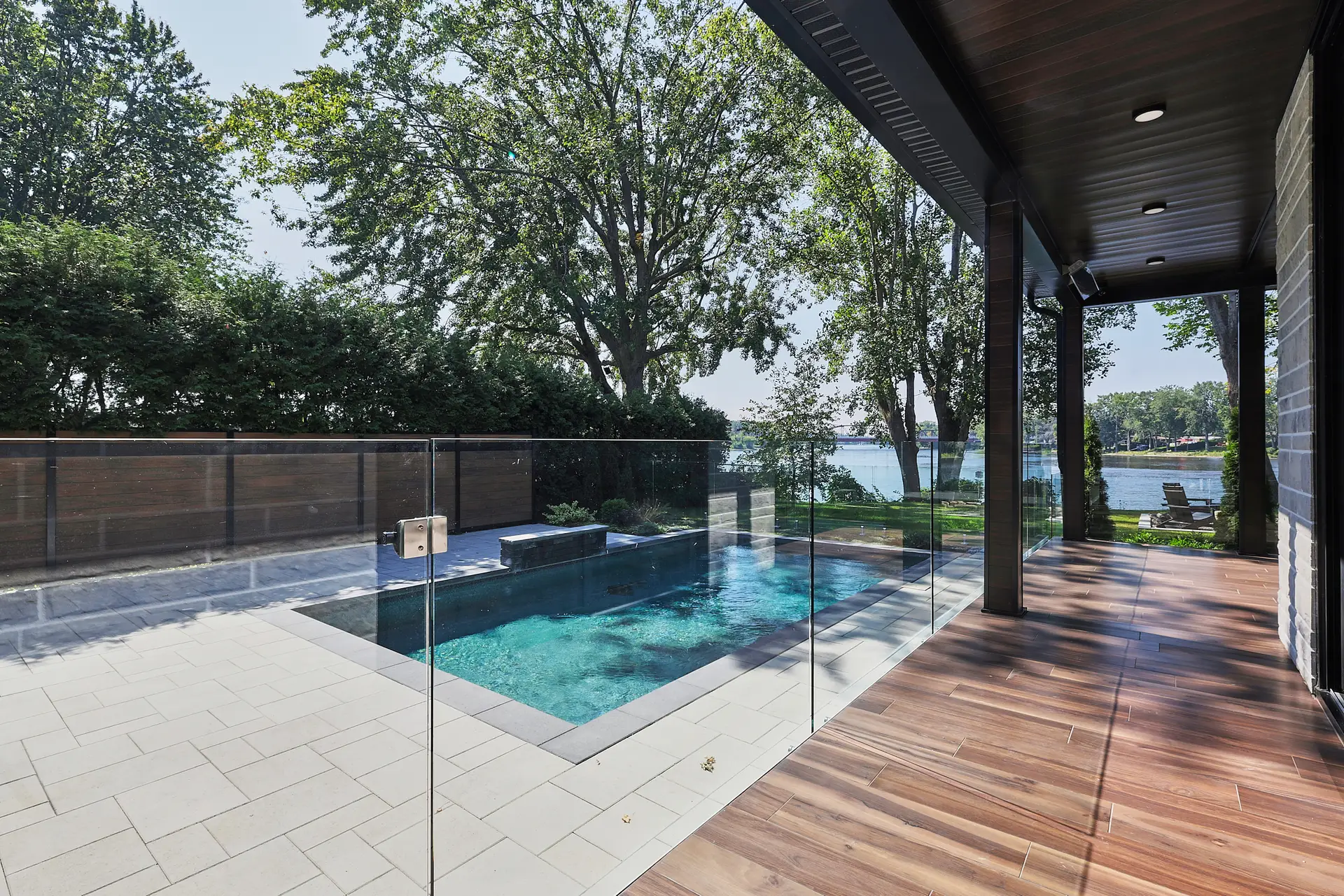 Inter Clôtures Sentinelle Modern patio with wood flooring overlooks a backyard pool surrounded by glass fencing, trees, and a view of a lake in the background. Fence Installation Quebec