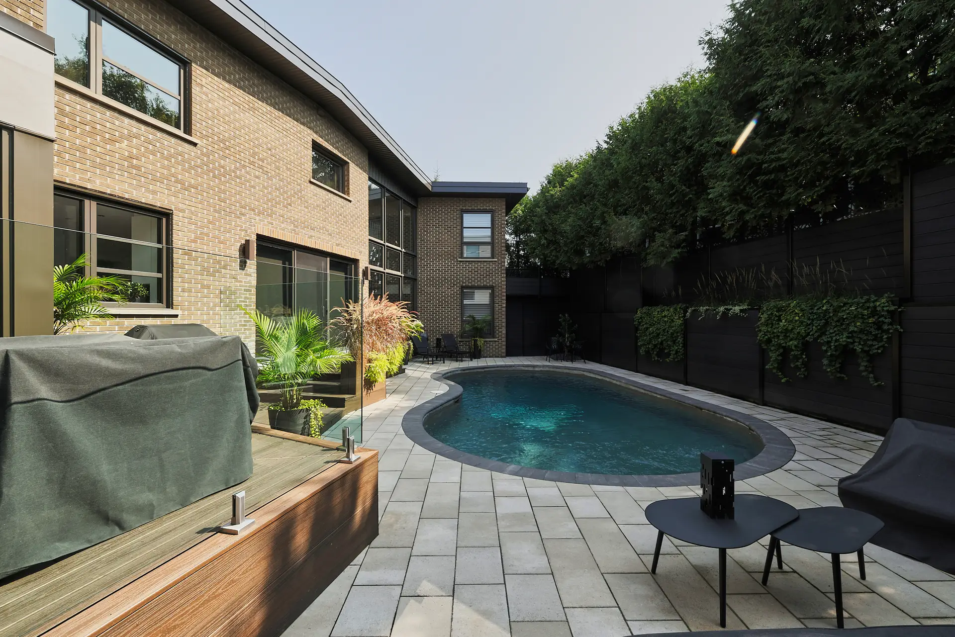 Inter Clôtures Sentinelle Modern backyard with a small swimming pool, patio seating area, potted plants, and a brick house with large windows. Fence Installation Quebec