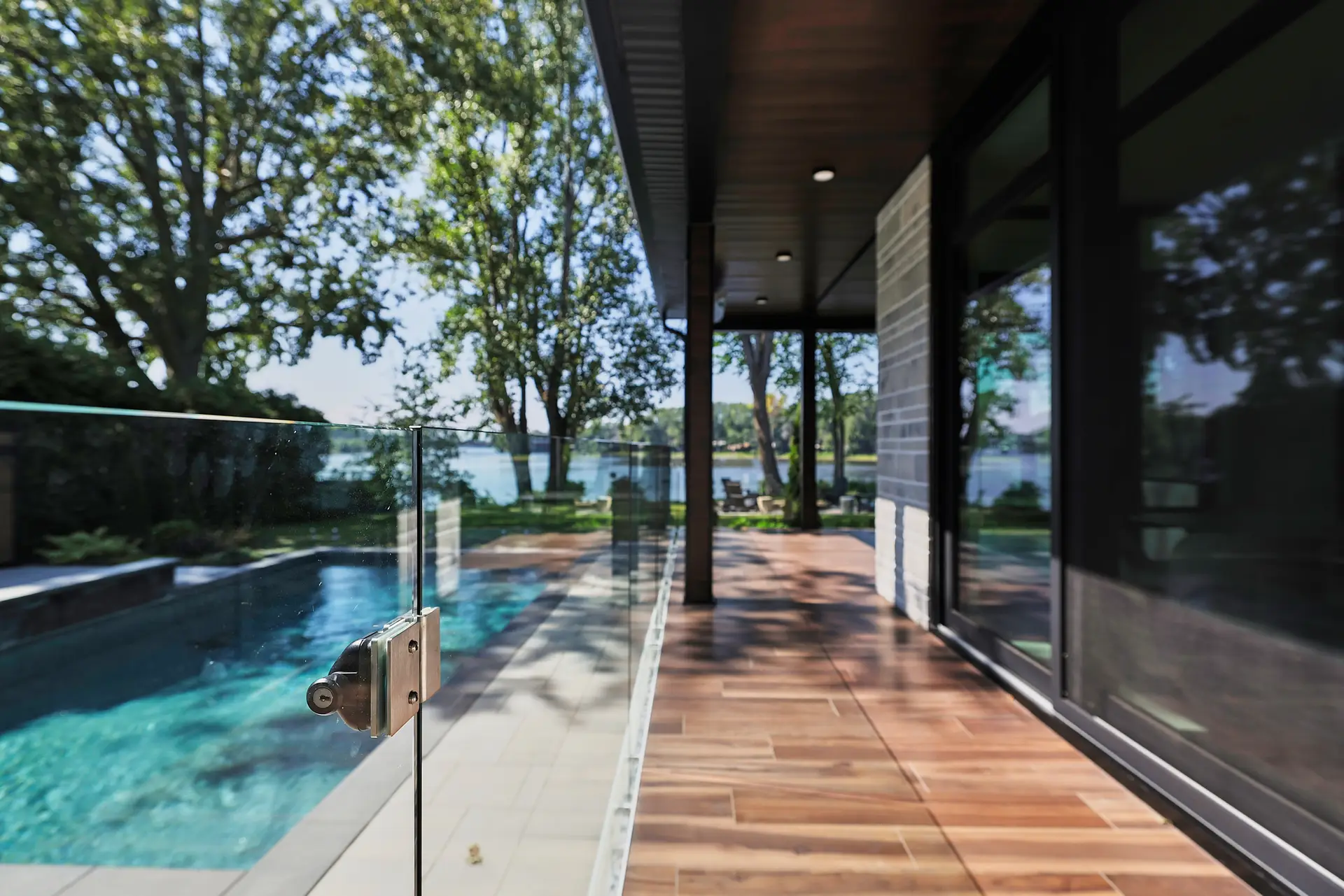 Inter Clôtures Sentinelle Modern house patio with wooden flooring, glass railing, and a view of an outdoor pool and trees near a lake under daylight. Fence Installation Quebec