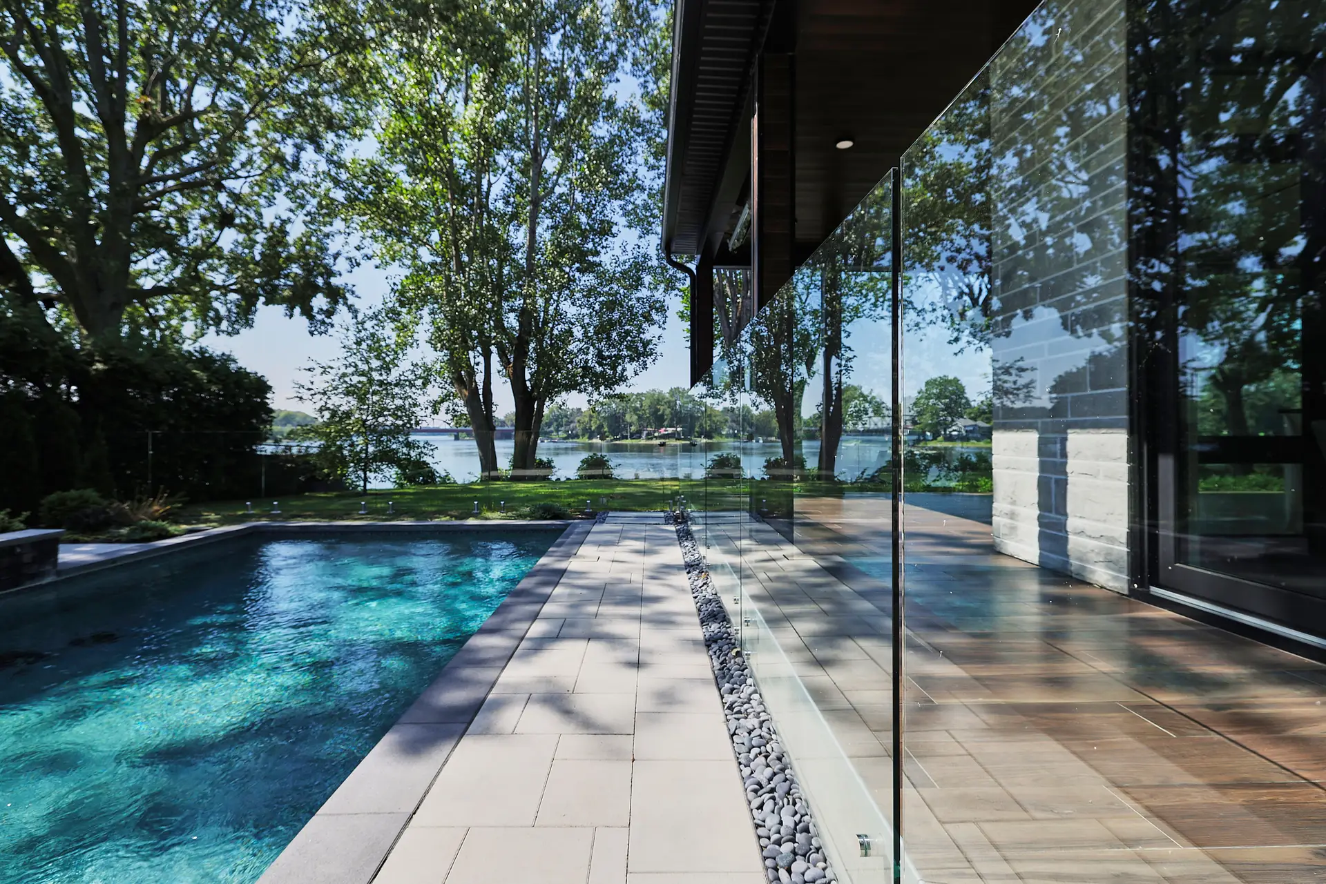 Inter Clôtures Sentinelle Modern house with glass walls beside a swimming pool, overlooking a lake and trees on a sunny day. Fence Installation Quebec