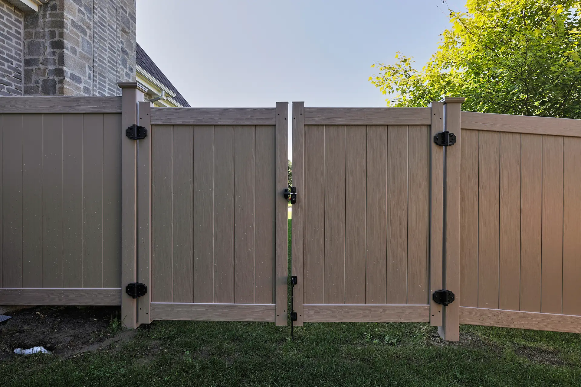 Inter Clôtures Sentinelle A closed tan vinyl privacy fence gate with black hinges stands on green grass, next to a brick house and leafy green trees under a clear sky. Fence Installation Quebec