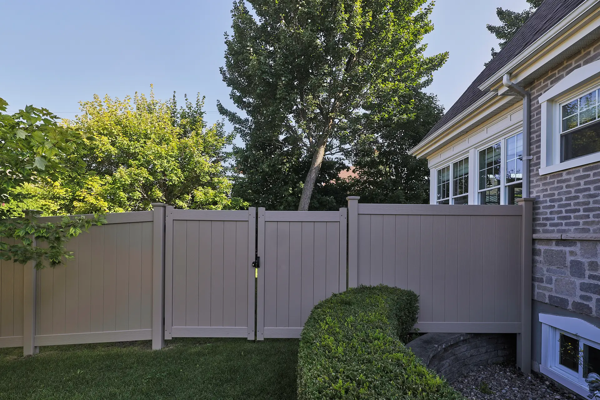 Inter Clôtures Sentinelle Beige vinyl privacy fence with a closed gate next to a brick house, green shrubbery in front, and tall leafy trees in the background under a clear blue sky. Fence Installation Quebec