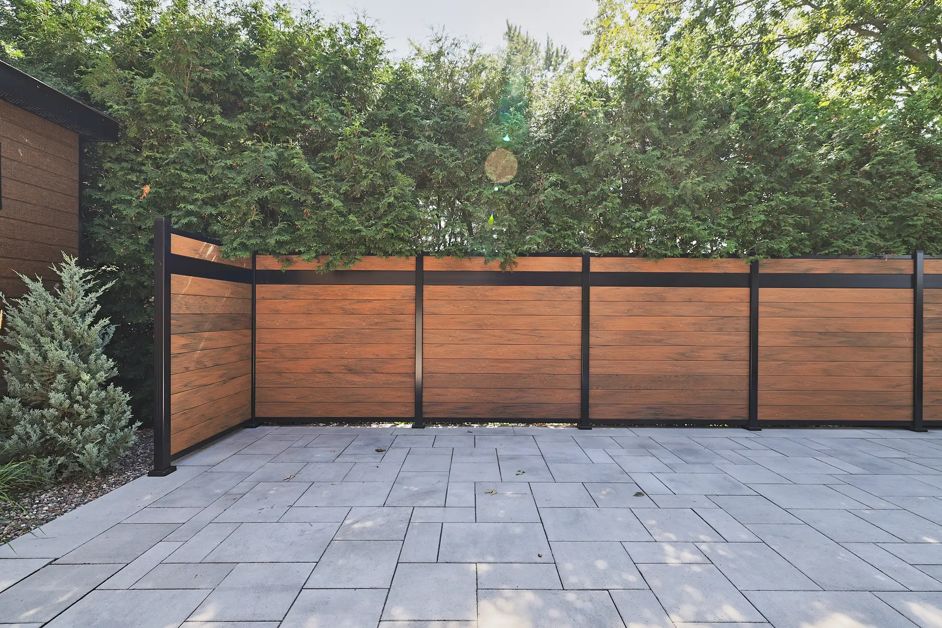Inter Clôtures Sentinelle Modern patio with large gray pavers, a horizontal wooden privacy fence with black posts, and tall green shrubs behind the fence. Fence Installation Quebec