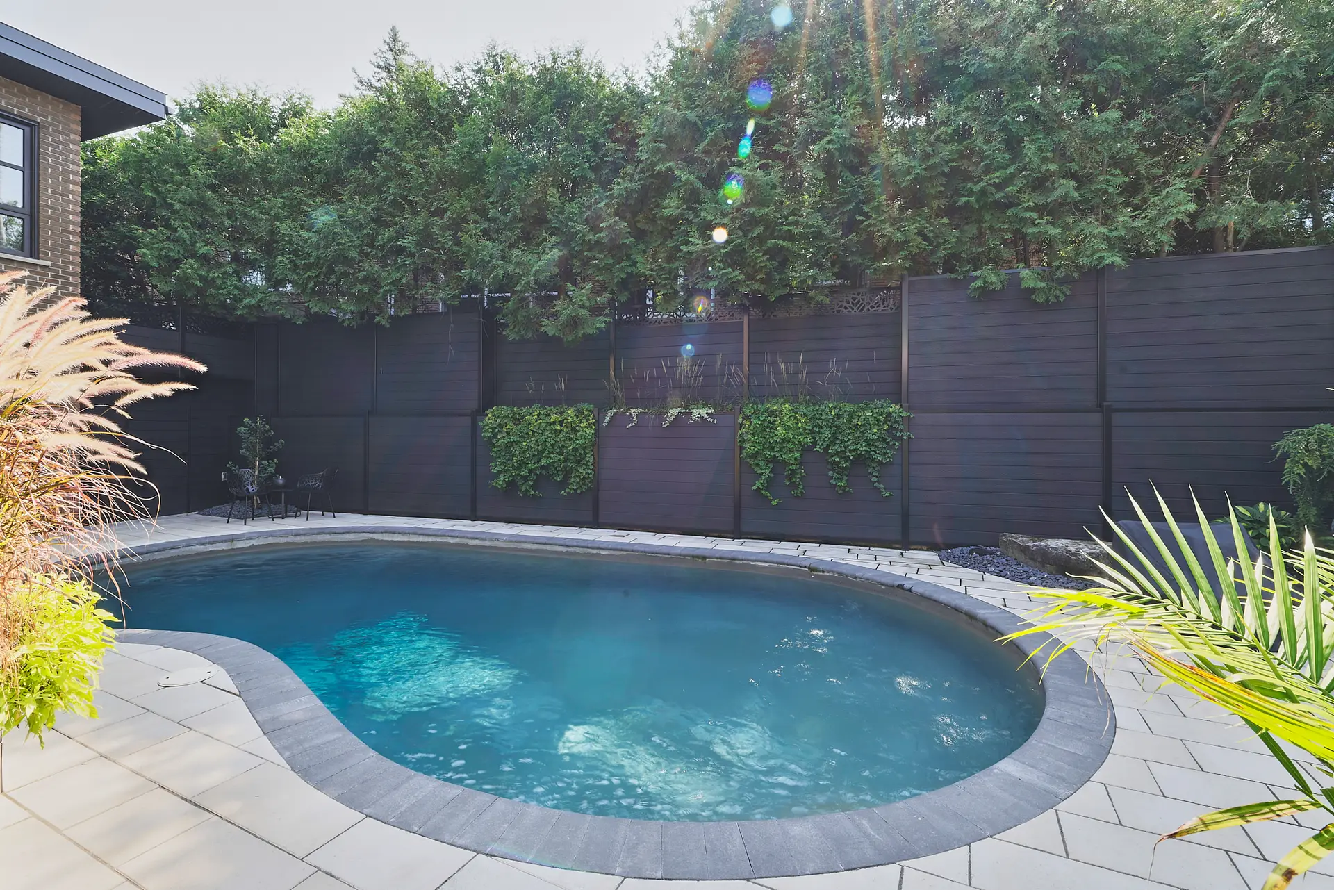 Inter Clôtures Sentinelle Backyard swimming pool with curved edges, stone tile deck, black privacy fence, green foliage, and sunlight streaming through trees. Fence Installation Quebec