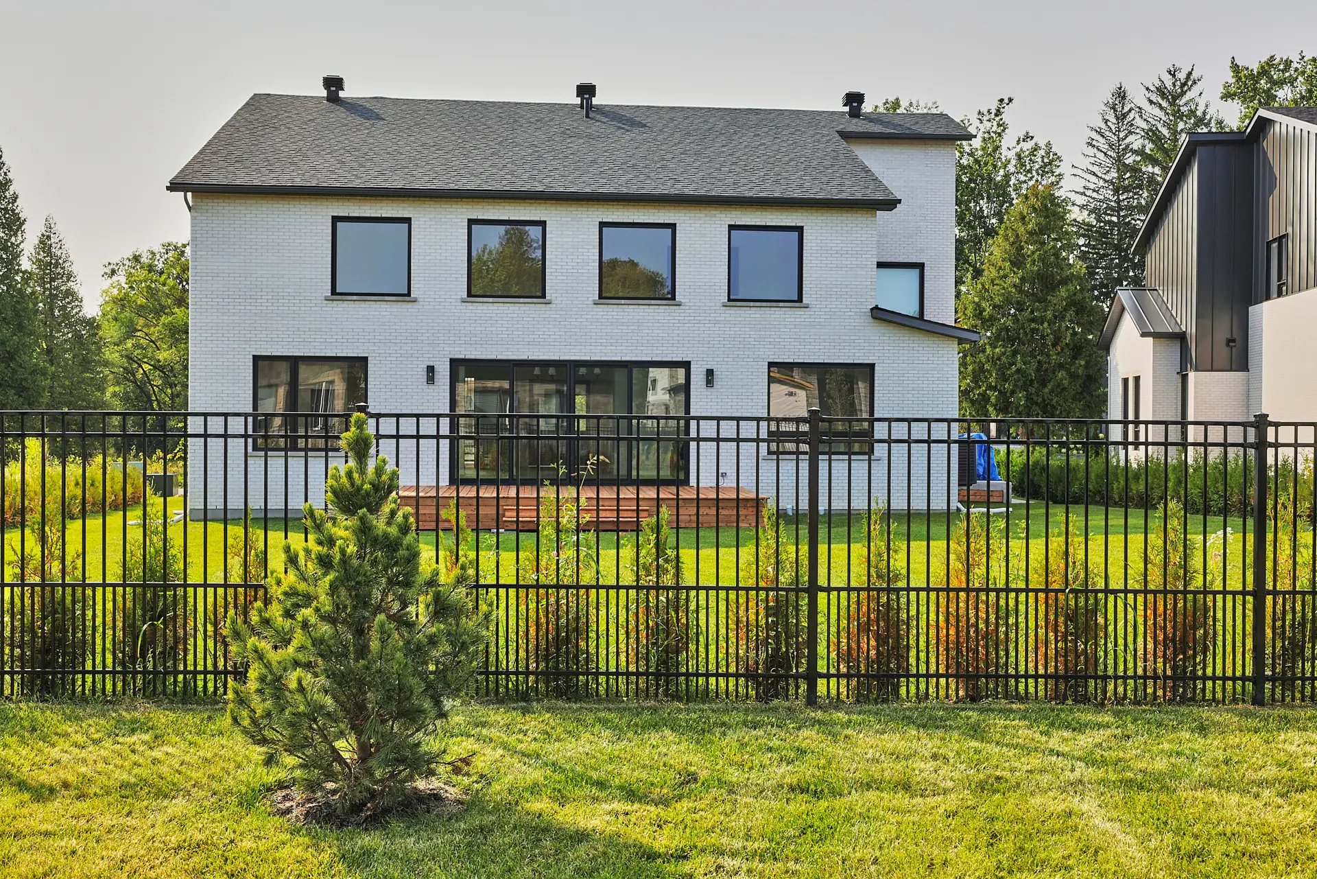 Inter Clôtures Sentinelle A modern two-story white house with large windows, a wooden deck, and a fenced backyard featuring grass, a small tree, and landscaping. Fence Installation Quebec