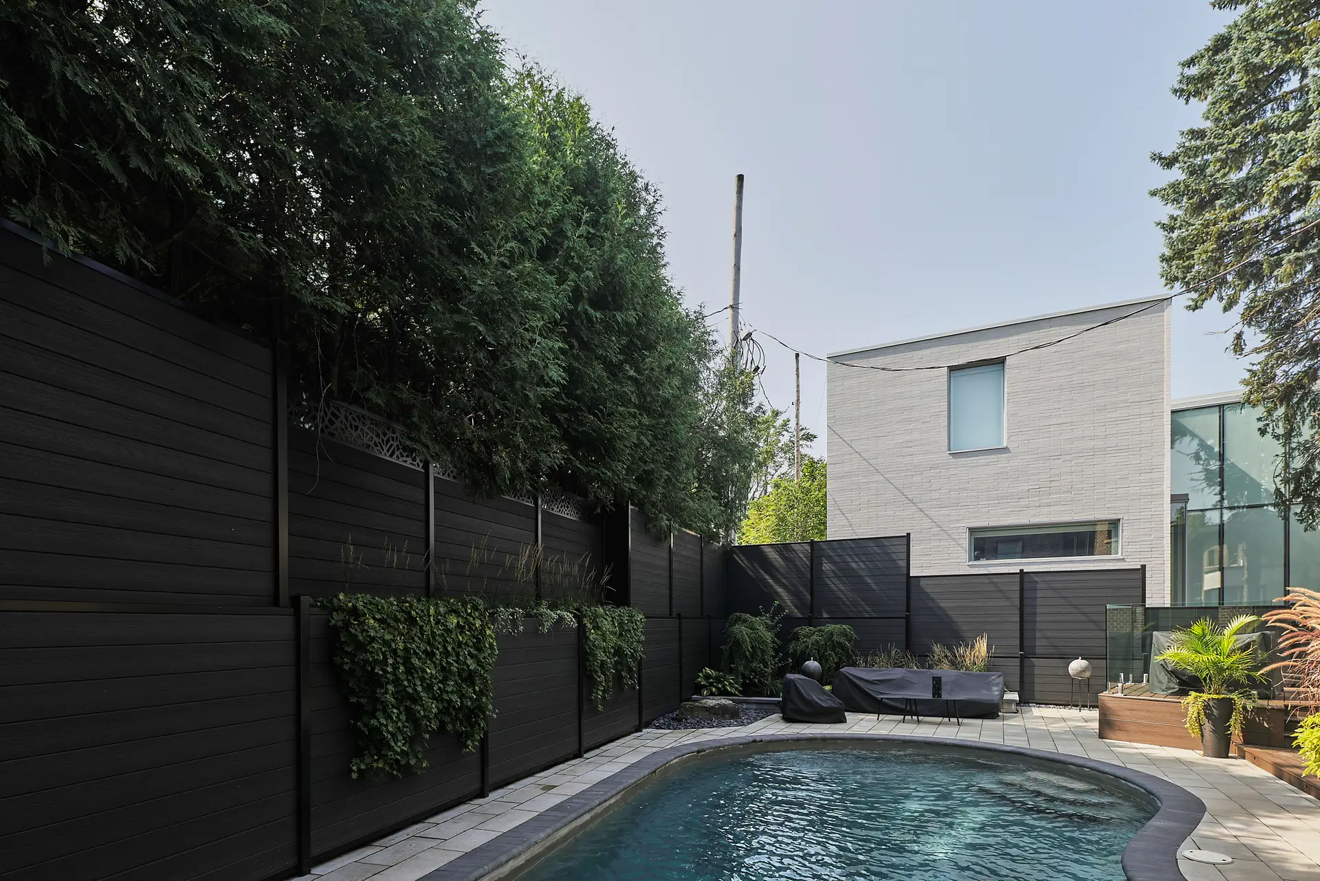 Inter Clôtures Sentinelle A backyard with a small curved pool, black privacy fence with greenery, patio seating, potted plants, and a modern gray house in the background. Fence Installation Quebec