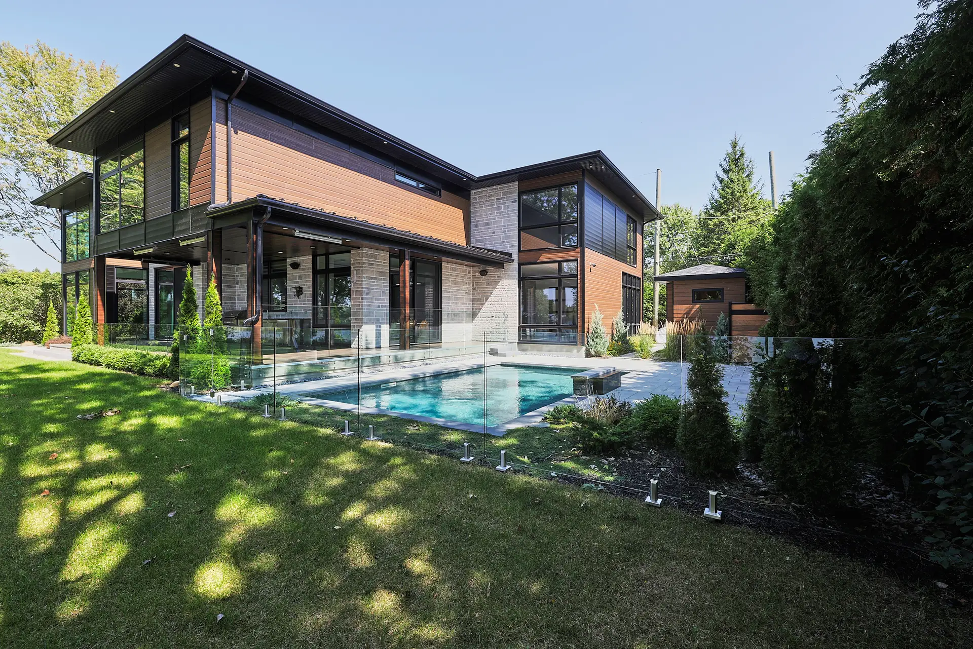 Inter Clôtures Sentinelle Modern two-story house with large windows, wood and stone exterior, glass pool fence, and a backyard swimming pool surrounded by grass and trees. Fence Installation Quebec