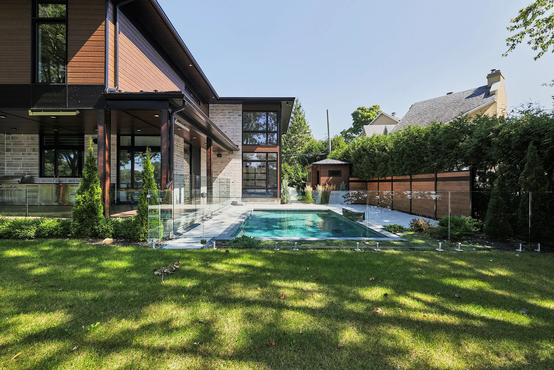 Inter Clôtures Sentinelle Modern house with large windows and wood accents, featuring a backyard swimming pool surrounded by glass fencing, green lawn, and tall hedges, on a sunny day. Fence Installation Quebec
