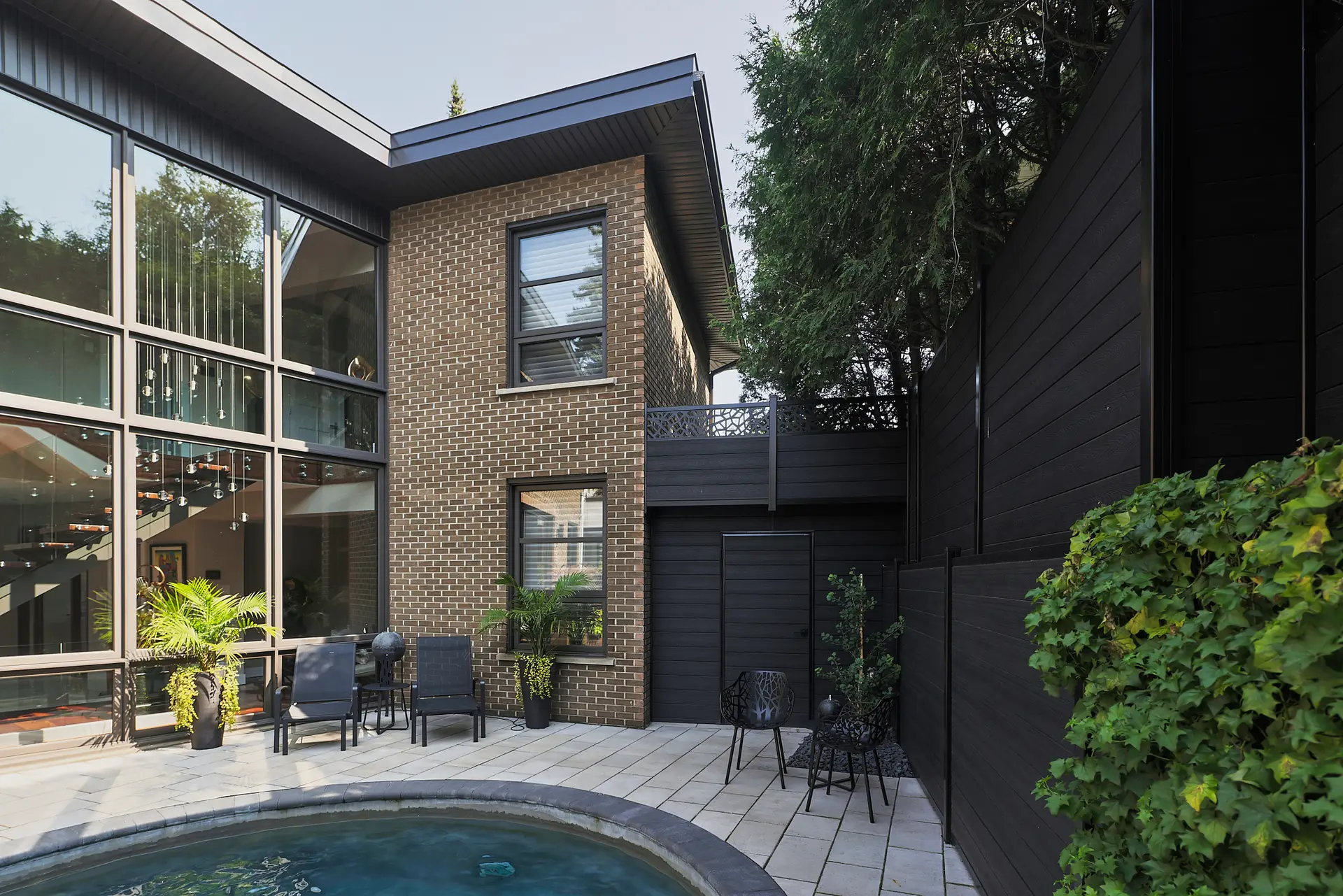 Inter Clôtures Sentinelle Modern brick house with large windows, patio seating, black privacy fence, and small round pool surrounded by plants and trees. Fence Installation Quebec