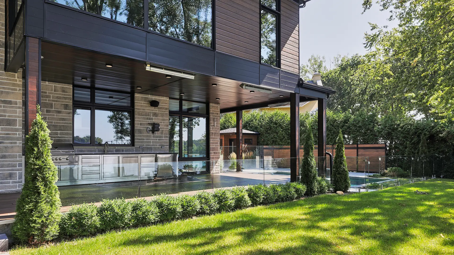 Inter Clôtures Sentinelle Modern backyard with covered patio, built-in outdoor kitchen, glass fencing, manicured hedges, and a swimming pool surrounded by trees and lawn. Fence Installation Quebec