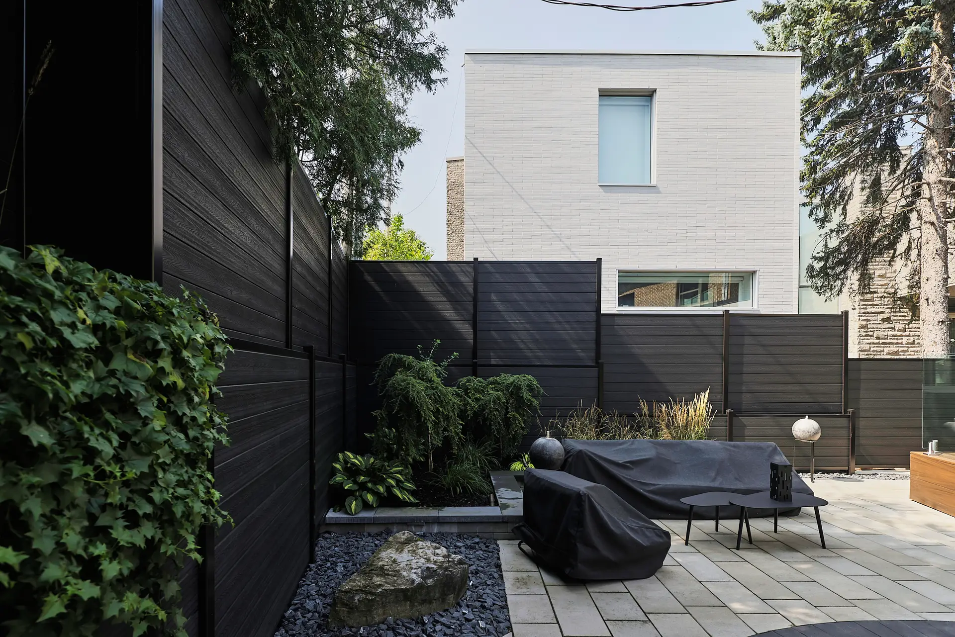Inter Clôtures Sentinelle Modern backyard with dark privacy fence, covered outdoor furniture, stone pavers, plants, and a large rock, with a gray two-story house in the background. Fence Installation Quebec